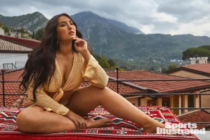 Yumi Nu sits on top of a red patterned cushion in front of the red rooftops in the city and the mountains beyond, wearing a tie-front yellow top.